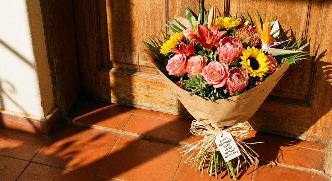 A vibrant bouquet of fresh birthday flowers wrapped in kraft paper resting on a sunlit doorstep in warm afternoon light.
